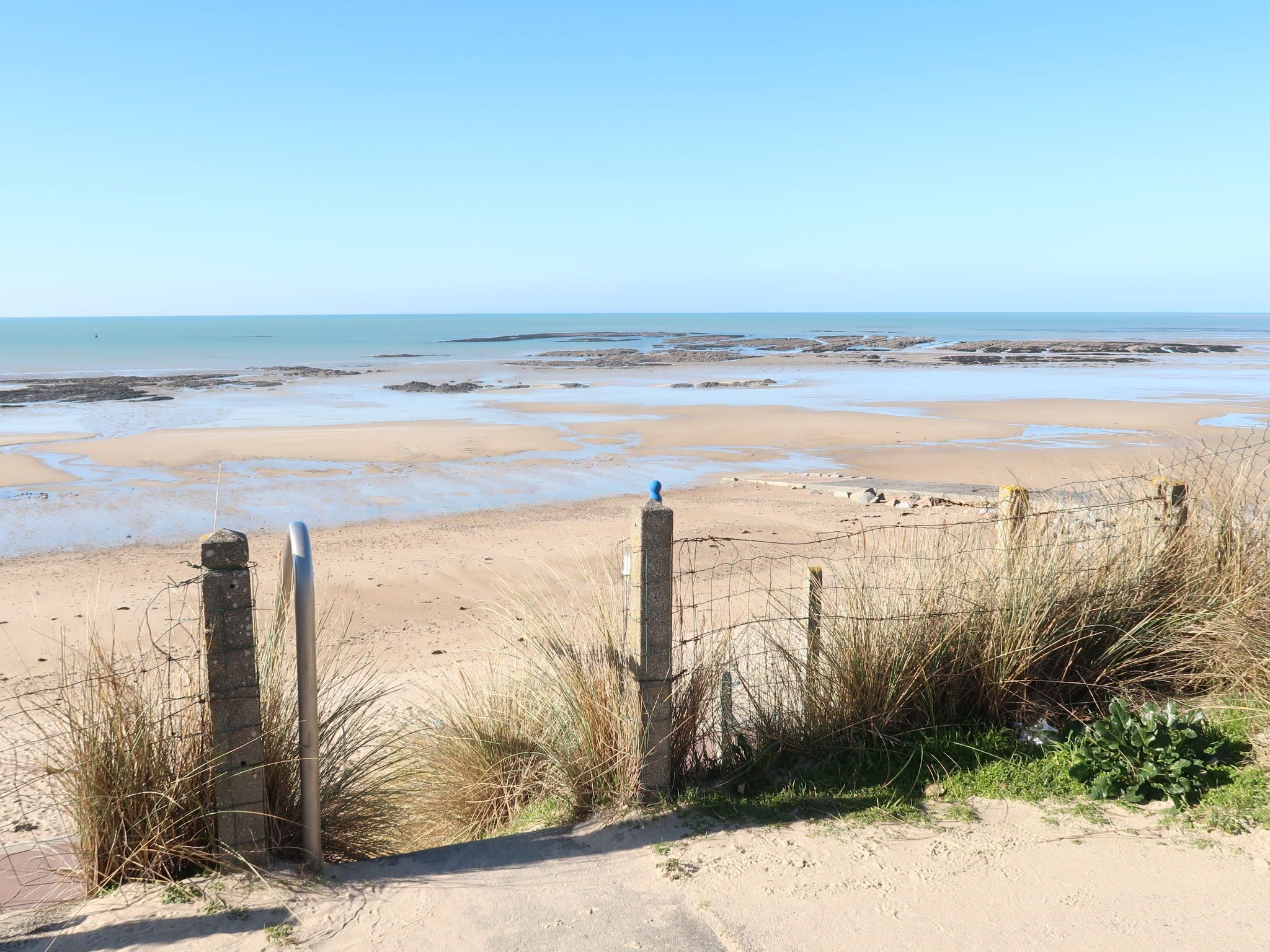 Ein Sandstrand mit freiem Blick auf das Meer, einer Holzlattenzaun und etwas Vegetation.