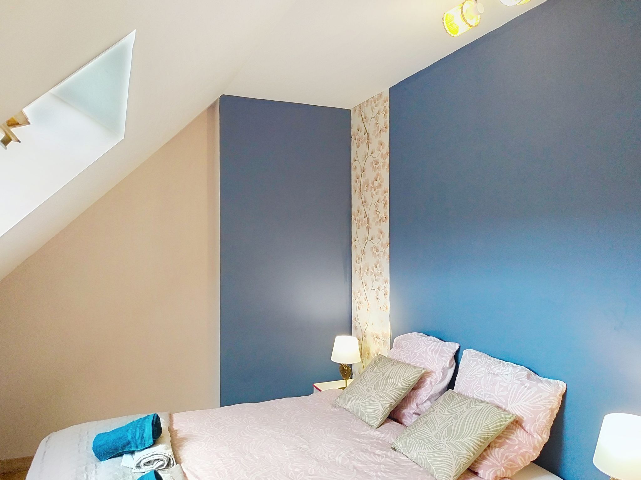 Een slaapkamer met schuine wanden, een bed met beddengoed met een patroon, twee bedlampen en een wand met bloemenbehang.