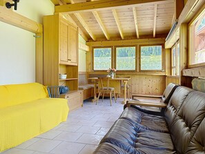 A cozy cabin interior with a wooden ceiling, a sofa, a dining table with chairs, and a window overlooking greenery.
