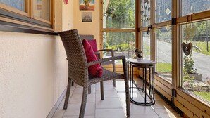 Ein gemütliches Zimmer mit Korbsessel, kleinem Tisch und Blick auf eine Straße und Grün durch große Fenster.
