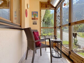 Ein gemütliches Zimmer mit Korbsessel, kleinem Tisch und Blick auf eine Straße und Grün durch große Fenster.