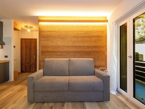 A modern living room with a grey sofa, wooden wall paneling, and a sliding glass door leading to an outdoor area. - Nordic Nest by Interhome (Bormio)