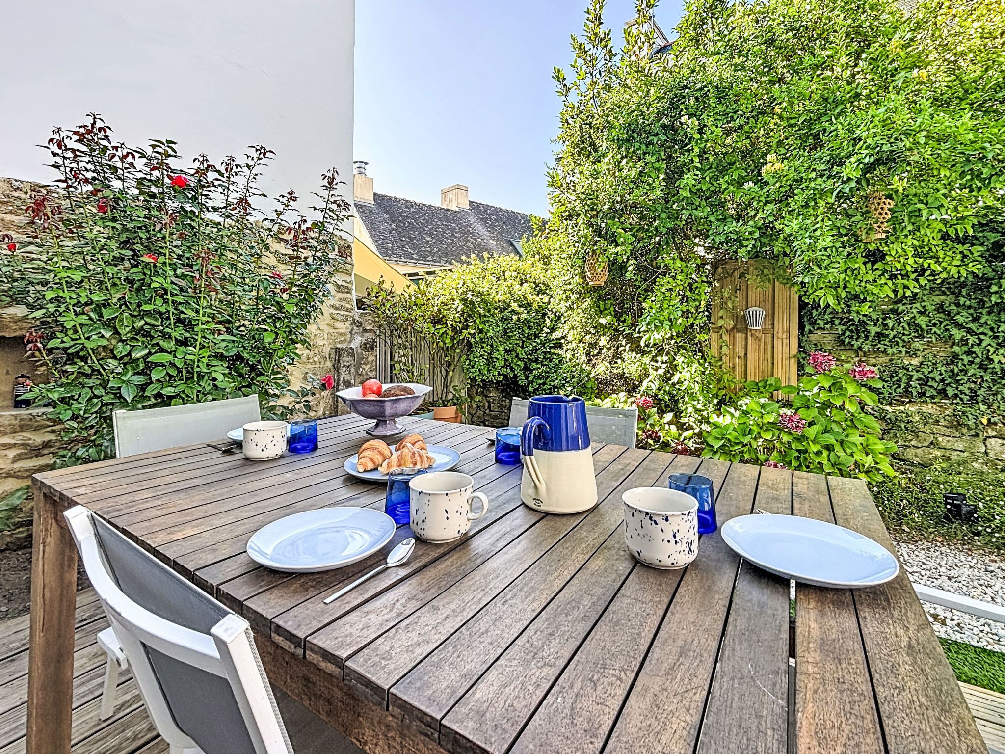 Une terrasse en bois avec une table dressée pour le petit-déjeuner, entourée d’une végétation luxuriante et d’un mur de pierre.