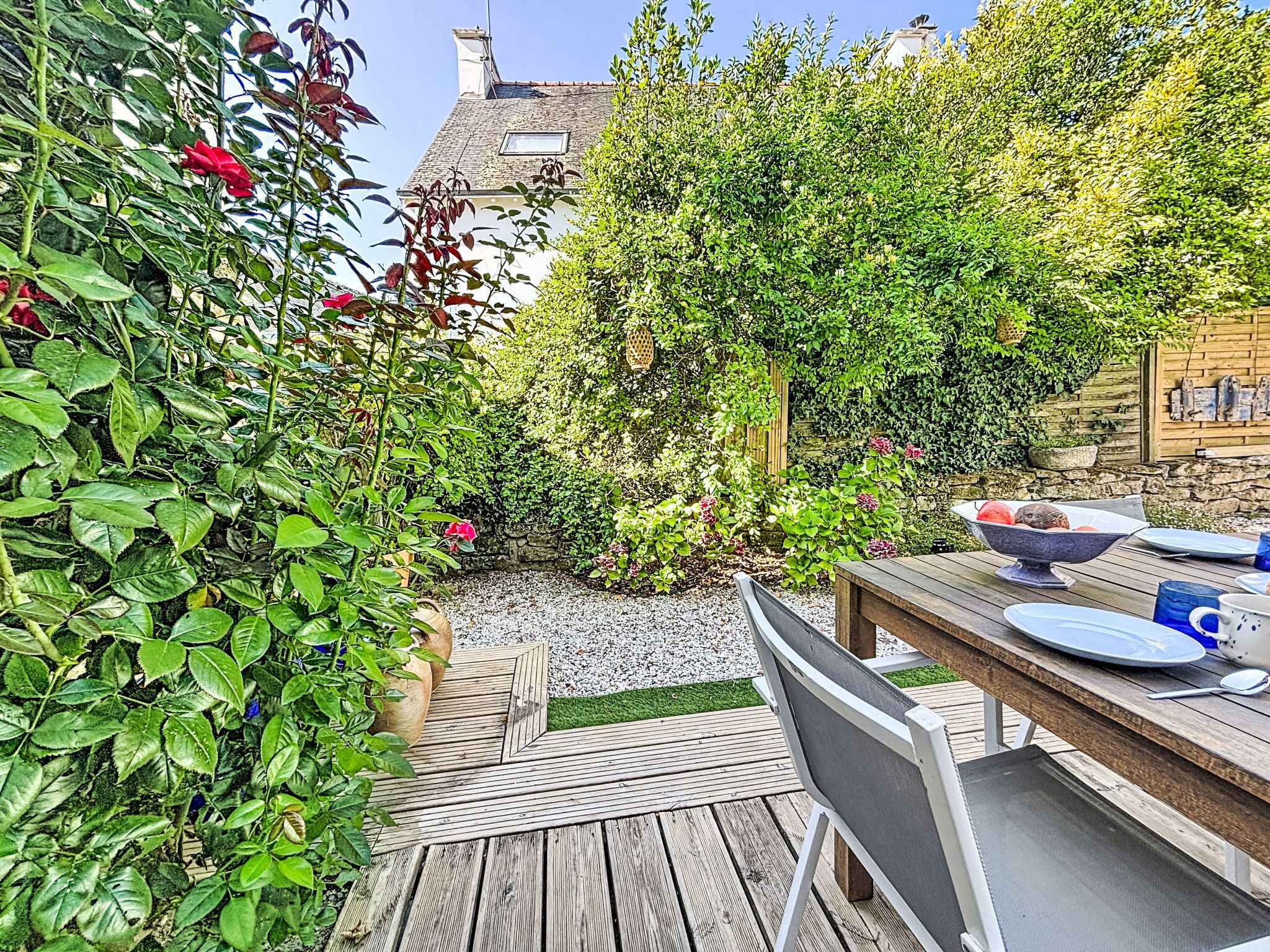 Eine Holzterrasse mit Tisch und Stühlen, umgeben von üppiger Bepflanzung und einer Steinsäule.