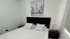 Een slaapkamer met een groot bed, een zwart hoofdeinde, wit beddengoed, een nachtkastje met een lamp, en een stadsgezichtschilderij aan de muur.
