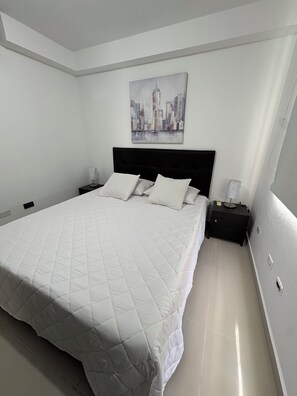A bedroom with a large bed, a black headboard, white bedding, a nightstand with a lamp, and a cityscape painting on the wall. - Amazing apartment. Prime, central location (Barranquilla)