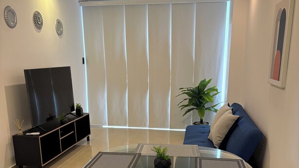 Een moderne woonkamer met een eettafel met glazen blad, grijze gestoffeerde stoelen, een zwarte tv-meubel en een groot raam met zonwering.