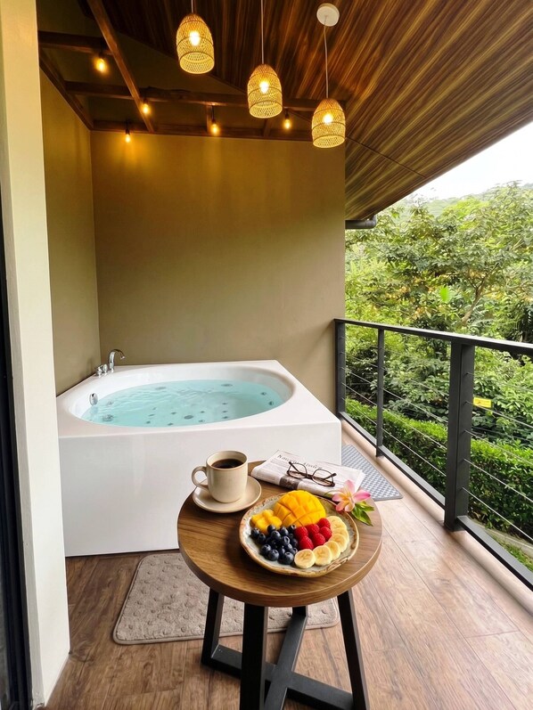 A balcony with a hot tub, a table with fruit and a cup of coffee, and a view of greenery.