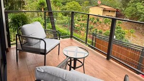 A balcony with a wooden ceiling, two chairs, a small round table, and a view of a green landscape with trees and a building. - Alto Bosque. Mountain chalet with a private hot tub ⛰️🌿 (Berlín)