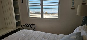 A bedroom with a bed, a window with a view of the beach, and a shelf unit.