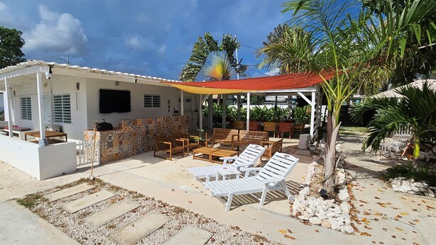 A covered outdoor seating area with lounge chairs and a dining table, surrounded by a white building with a tiled roof and a patterned wall. - Private Beachfront Estate w/ Infinity Pool (Santa Isabel)