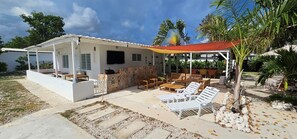 A covered outdoor seating area with lounge chairs and a dining table, surrounded by a white building with a tiled roof and a patterned wall.