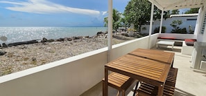 A balcony with a wooden table and bench overlooking a rocky beach and the sea.