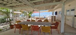 A covered outdoor seating area with a wooden table and colorful plastic chairs, overlooking a beach.