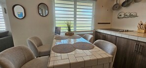 A dining area with a tiled table, four chairs, a potted plant, and a kitchen with cabinets and utensils.