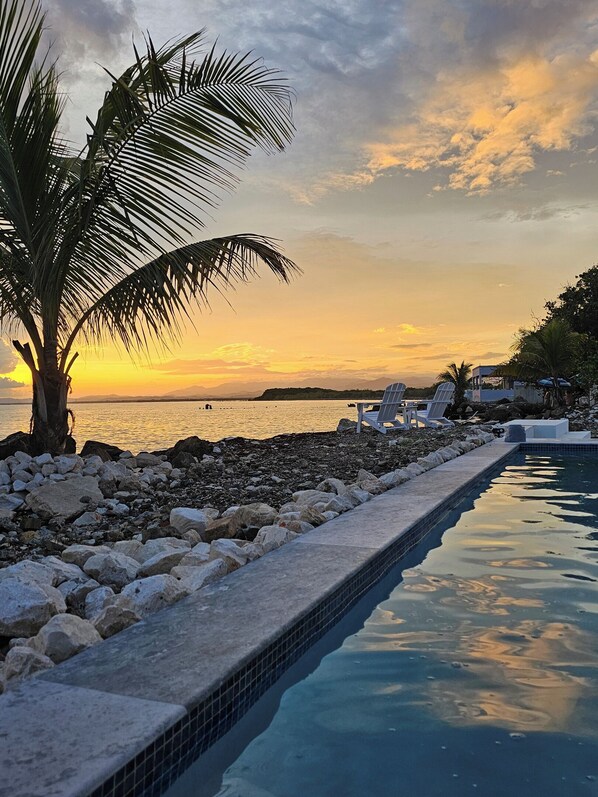 A poolside area with a view of the sunset over the water, surrounded by rocks and palm trees. - Private Beachfront Estate w/ Infinity Pool (Santa Isabel)