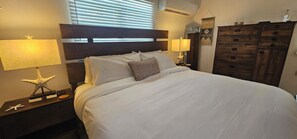 A neatly made bed with white linens, a wooden headboard, and a decorative pillow. There are two bedside tables with lamps and starfish decorations. A wooden dresser with drawers is to the right.