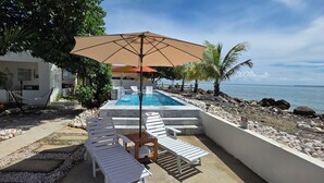 A pool area with lounge chairs and a large umbrella, surrounded by a rocky shoreline and palm trees. - Private Beachfront Estate w/ Infinity Pool (Santa Isabel)