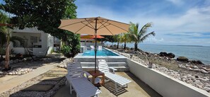 A pool area with lounge chairs and a large umbrella, surrounded by a rocky shoreline and palm trees. - Private Beachfront Estate w/ Infinity Pool (Santa Isabel)