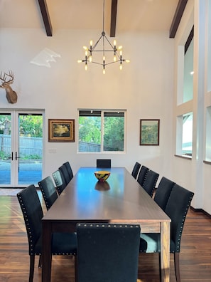 A dining area with a long table, six chairs, a chandelier, and two windows with views of greenery.