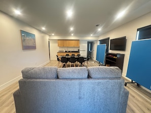 Ein modernes Wohnzimmer mit einem grauen Sofa, einem Essbereich mit Tisch und Stühlen, einer Küche im Hintergrund und einem an der Wand befestigten Fernseher.