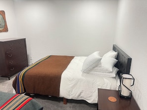 A neatly made bed with white linens and a brown throw, a wooden nightstand with a lamp, and a dark wooden dresser against a white wall.