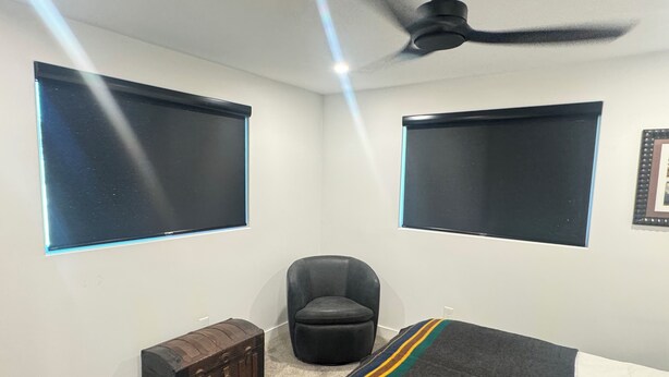 Une chambre avec deux grandes fenêtres, un ventilateur de plafond, une chaise, une petite commode en bois et un lit recouvert d’une couverture à rayures.