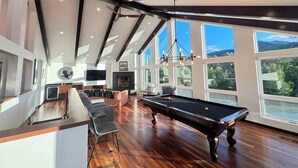 Un vaste espace de vie comprenant une table de billard, des sièges et de grandes fenêtres offrant une vue sur l’extérieur.