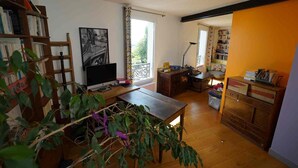 Et værelse med et træskrivebord, en computer, en reol, et vindue med gardiner og en plante.