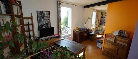 A room with a wooden desk, a computer, a bookshelf, a window with curtains, and a plant.