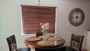 Un coin repas comprenant une table ronde, quatre chaises, un vase de fleurs, une horloge murale et une fenêtre avec des stores.