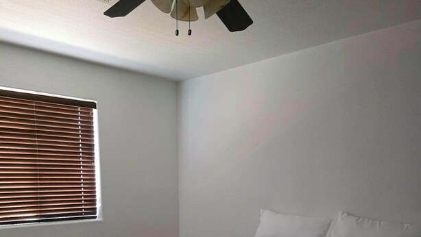 Une chambre à coucher avec un lit, un ventilateur de plafond, une fenêtre avec des stores et un sol recouvert de moquette.