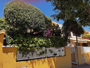 En gul bygning med et hvidt espalier, et træ og lyserøde blomster.