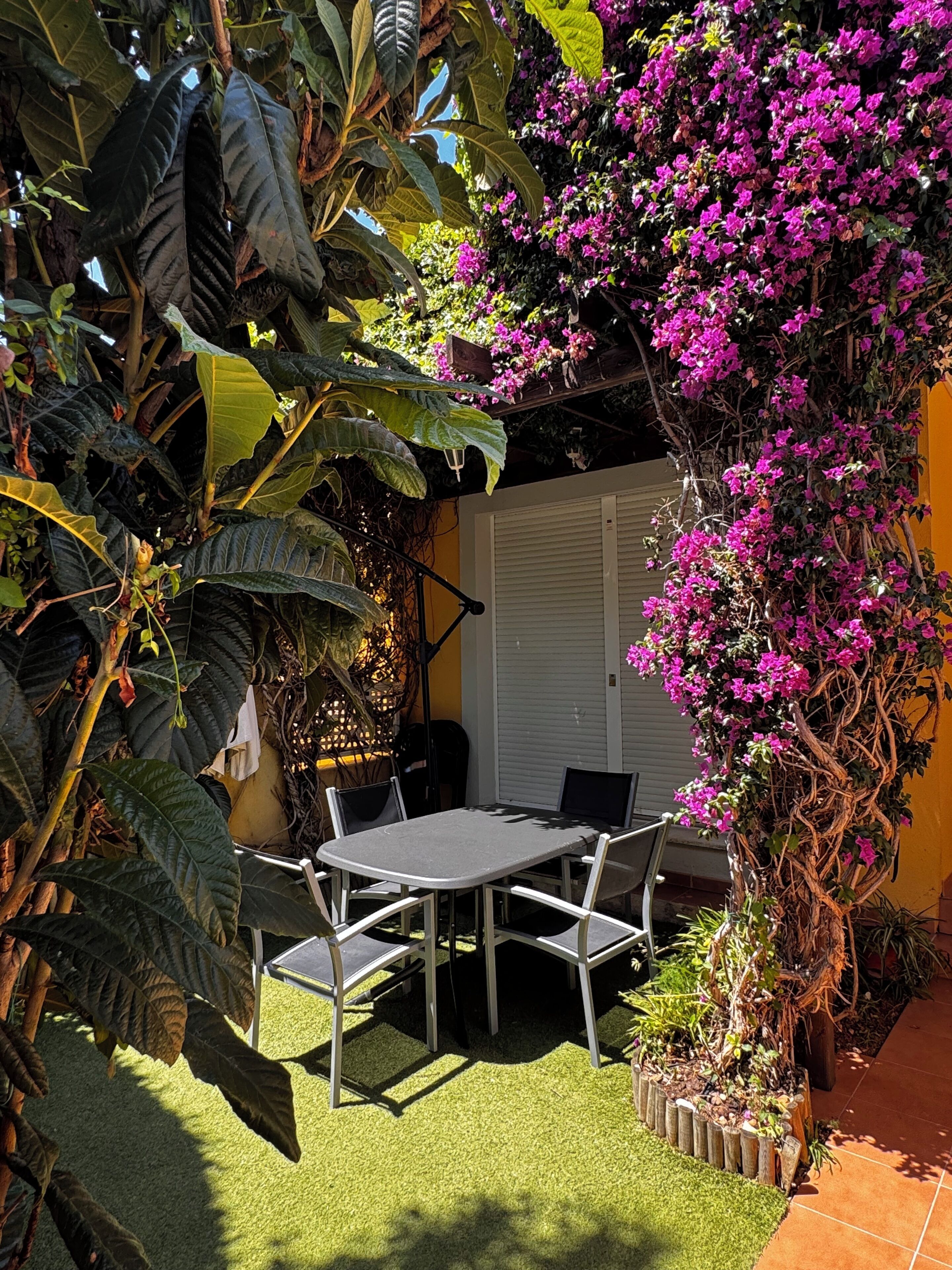 A patio with a table and chairs, surrounded by lush greenery and flowering plants.