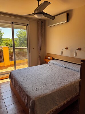 A bedroom with a bed, a ceiling fan, an air conditioning unit, a window with a view of greenery, and a balcony with a yellow railing. - 4-bedroom house in a gated community with a pool and beaches just 5 minutes away (Alcalà de Xivert)