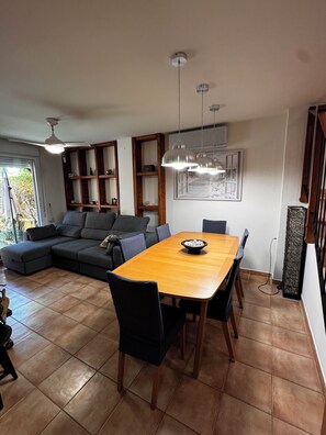 A modern living room with a sectional sofa, a wooden dining table with chairs, pendant lights, and a large window. - 4-bedroom house in a gated community with a pool and beaches just 5 minutes away (Alcalà de Xivert)