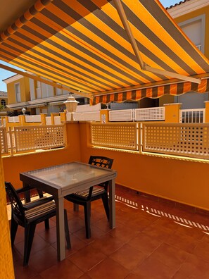 An outdoor patio with a table and chairs, a striped awning, and a view of a yellow building. - 4-bedroom house in a gated community with a pool and beaches just 5 minutes away (Alcalà de Xivert)