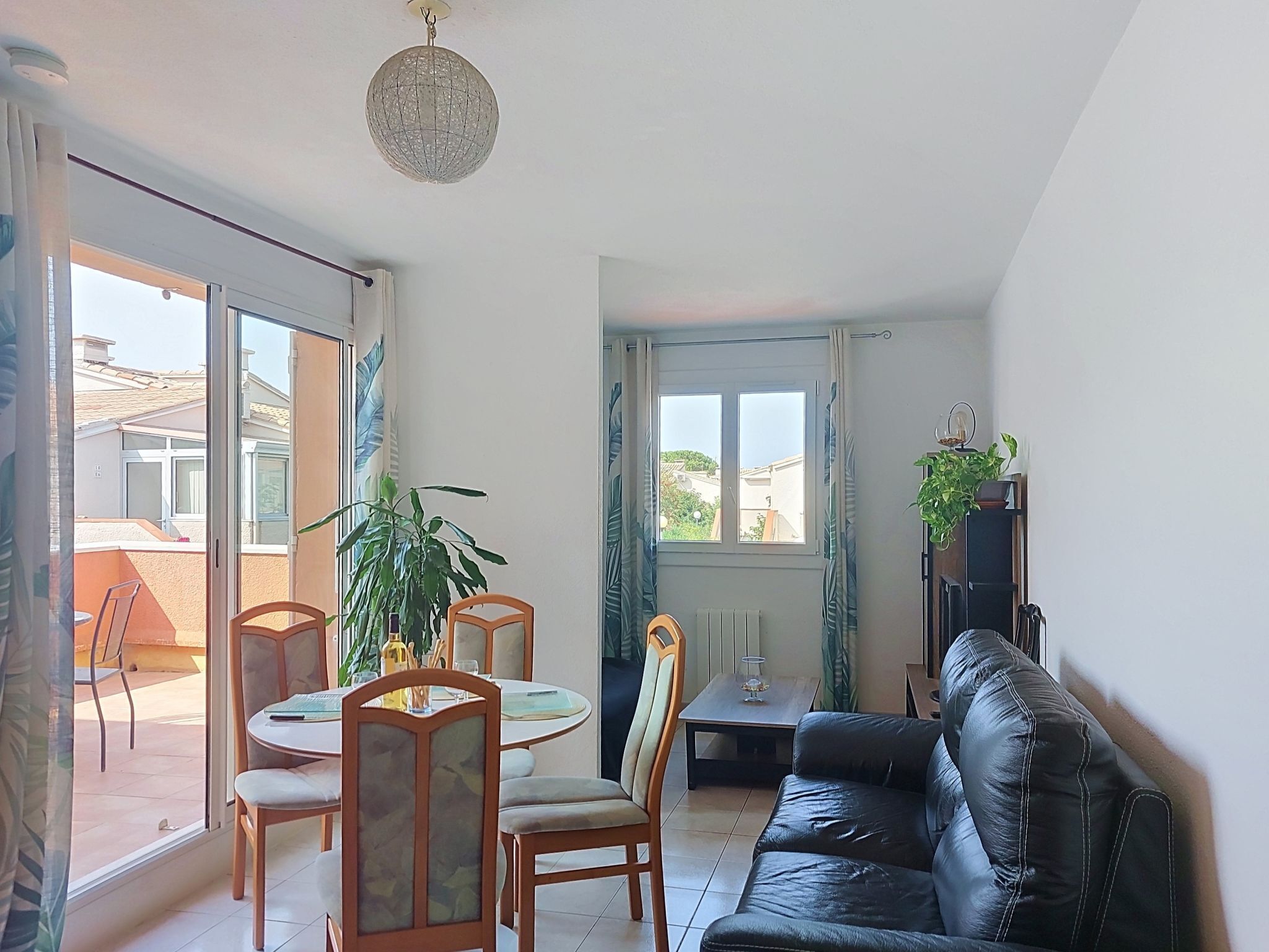 A living room with a black leather sofa, a round dining table with chairs, a potted plant, and a ceiling light fixture.