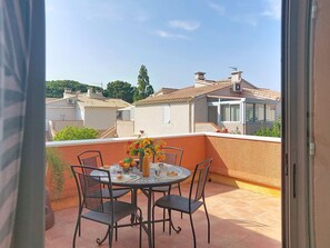 Un balcon avec une table dressée pour deux personnes, entouré de chaises, donnant sur un quartier résidentiel avec des maisons et des arbres.