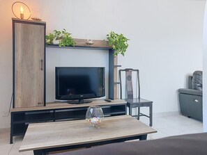 A living room with a flat-screen TV on a wooden stand, a glass coffee table with a decorative item, and a grey sofa. - Les Nouvelles Mirandas by Interhome (Saint Cyprien)
