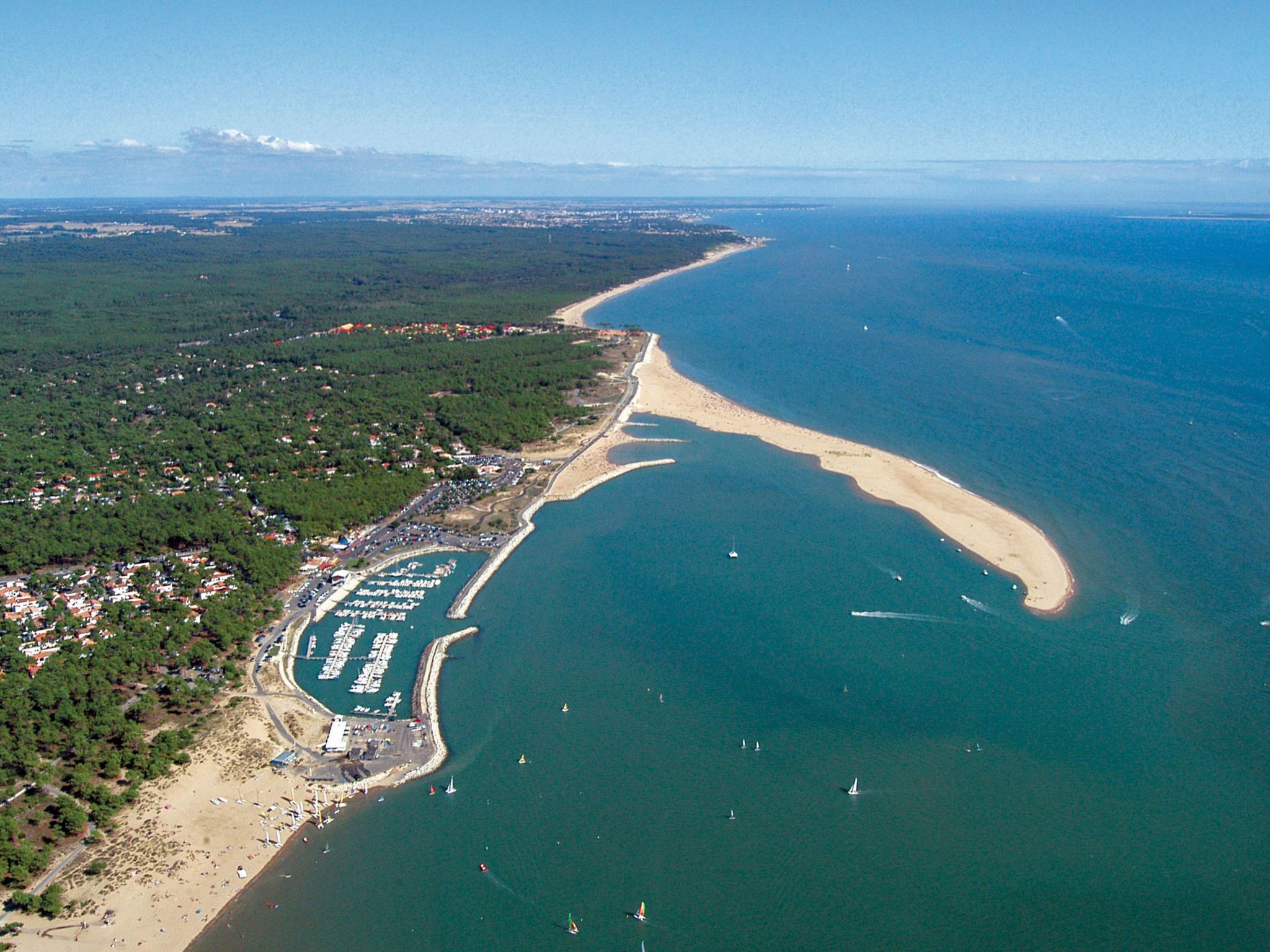 Vue aérienne d’une zone côtière avec une plage, un port de plaisance et des immeubles résidentiels.