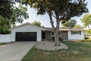 A single-story house with a garage, a tree in front, and a well-maintained lawn. - 4 BR, 3 bath ranch home featuring 2 King & 2 Queen bedrooms  (Sioux Falls)