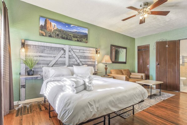 A bedroom with a bed, a ceiling fan, a nightstand, a lamp, a chair, and a wooden floor.