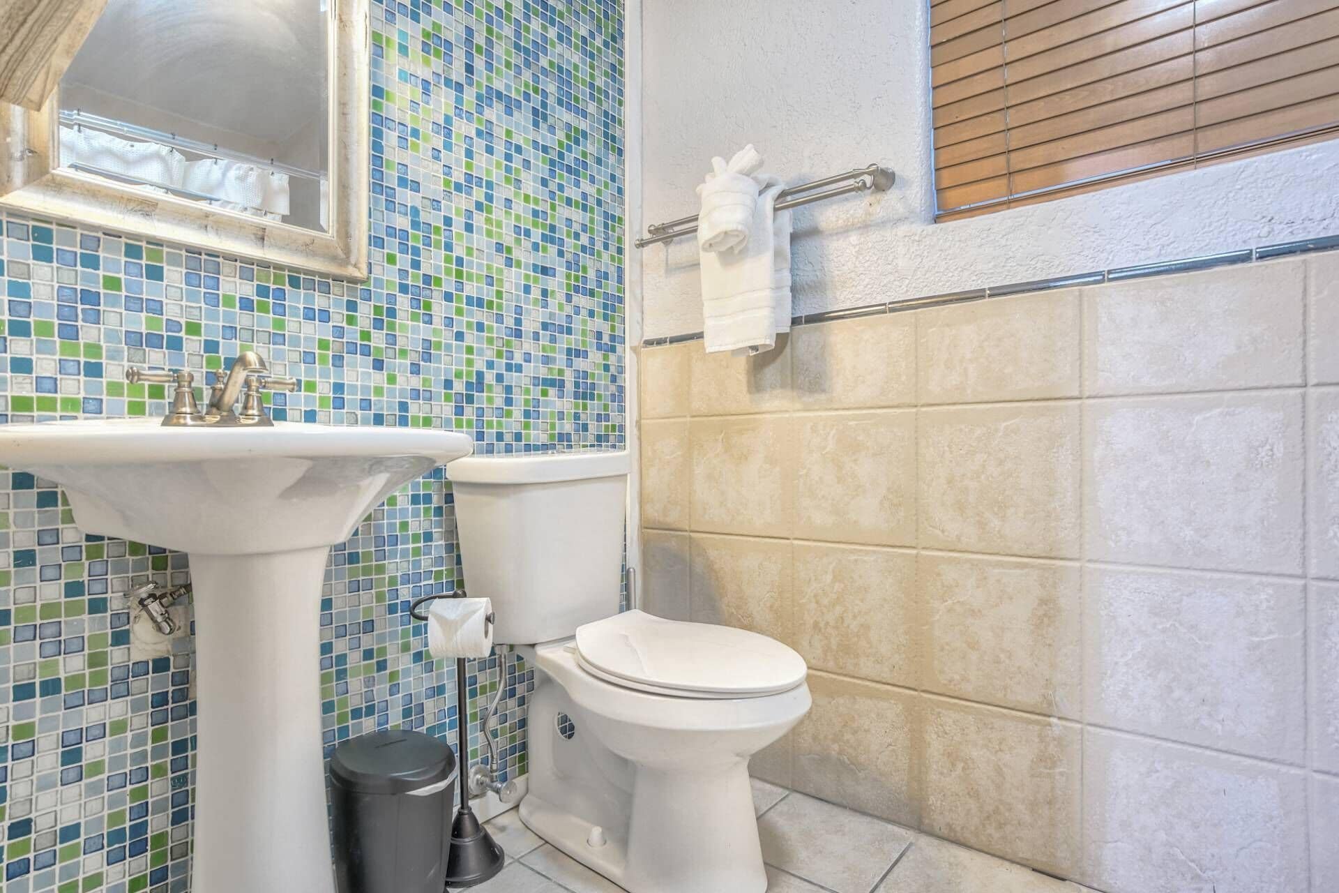 Un baño con un salpicadero de mosaicos, un lavamanos pedestal, un inodoro y un cubo de basura.