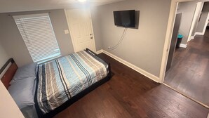 Quarto com cama, televisão presa à parede, porta e janela com persianas.