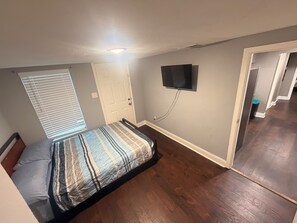 Ein Schlafzimmer mit einem Bett, einem an der Wand montierten Fernseher, einer Tür und einem Fenster mit Jalousien.