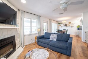 A living room with a fireplace, a sofa, a coffee table, and a dining area with chairs and a table. - Eikos Beach House - 4 BR / 3 BA - Heated Pool / Spa (Treasure Island)