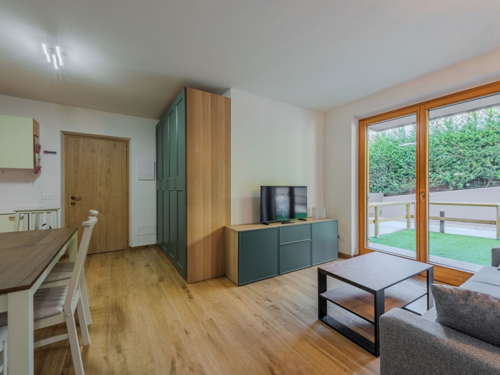 A modern living room with a wooden dining table, a sofa, a TV on a cabinet, and a sliding glass door leading to a garden.