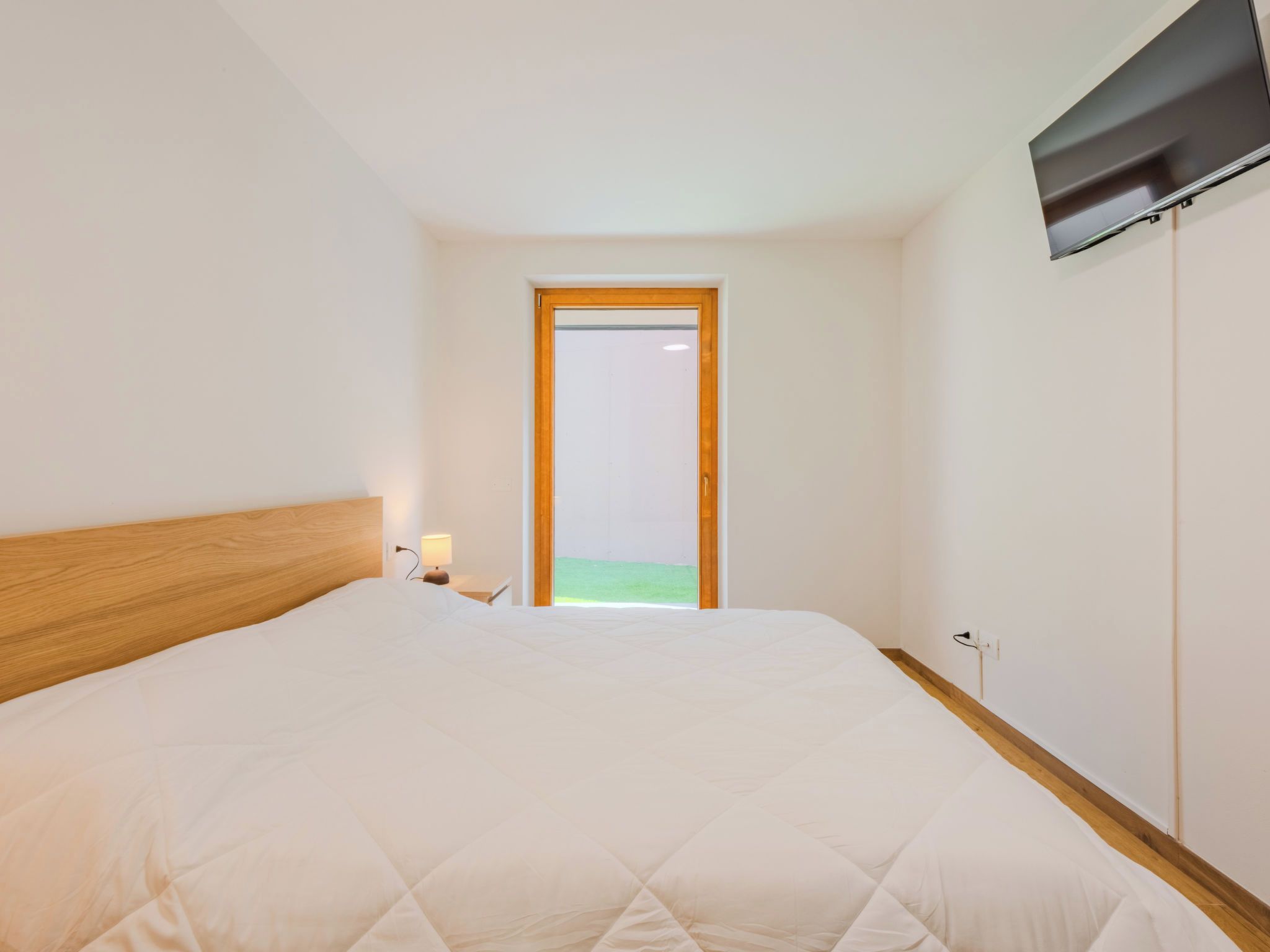 A bedroom with a large bed, a nightstand with a lamp, a window with a view of greenery, and a wall-mounted TV.