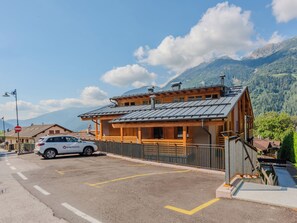Un edificio de madera de dos pisos con techo inclinado, un coche estacionado al frente y montañas al fondo.